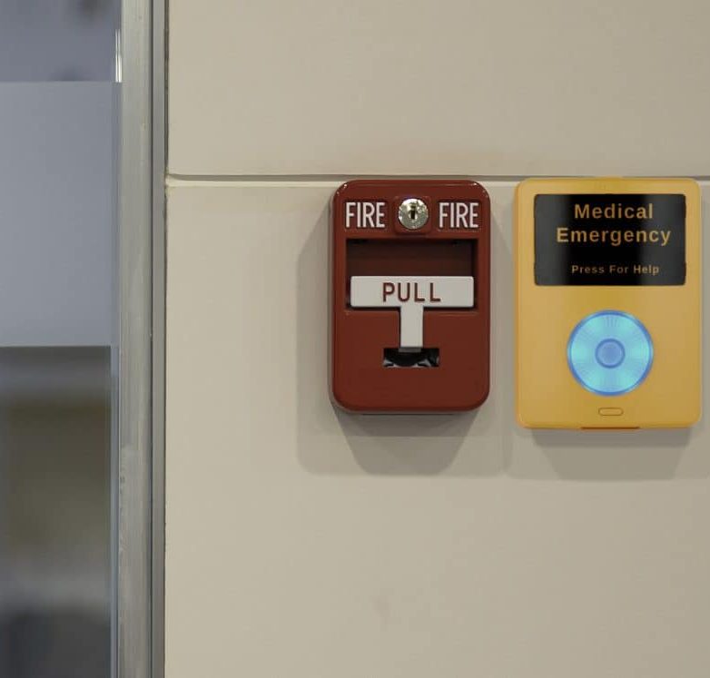 An image of an Algo 1202 call button securely mounted on a wall for emergency notification. The product is yellow with text that reads medical emergency, press for help and a big circular button that is glowing in blue. It is positioned at an accessible height next to a fire alarm.
