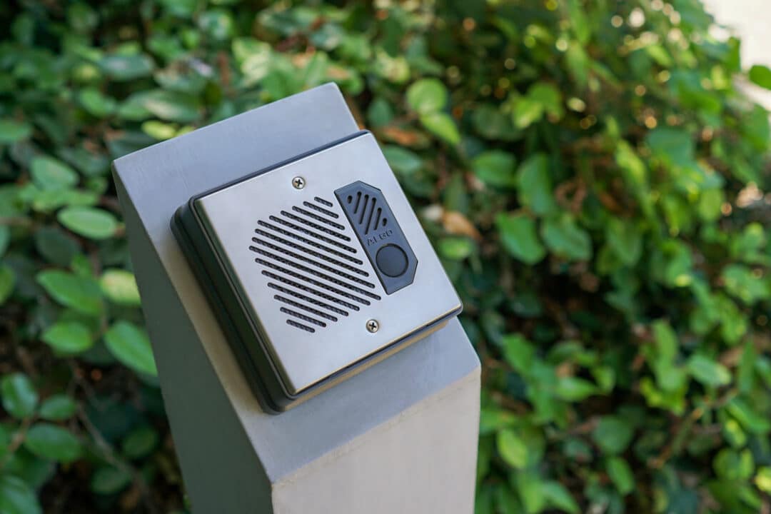 An image of an Algo 8201 door intercom system installed outside a building. The intercom unit is securely mounted on a post near the entrance. It features a speaker, microphone, and call button to initiate or accept intercom calls.